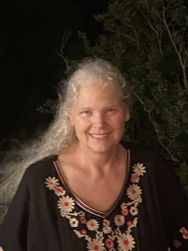 Smiling woman with long gray hair, wearing a black embroidered top, outdoors at night.